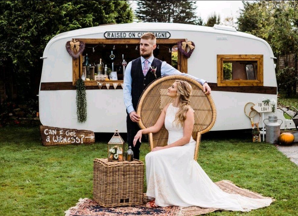 Bride and groom pose near a vintage caravan bar in a garden. Bride sits in a peacock chair; groom stands behind her.