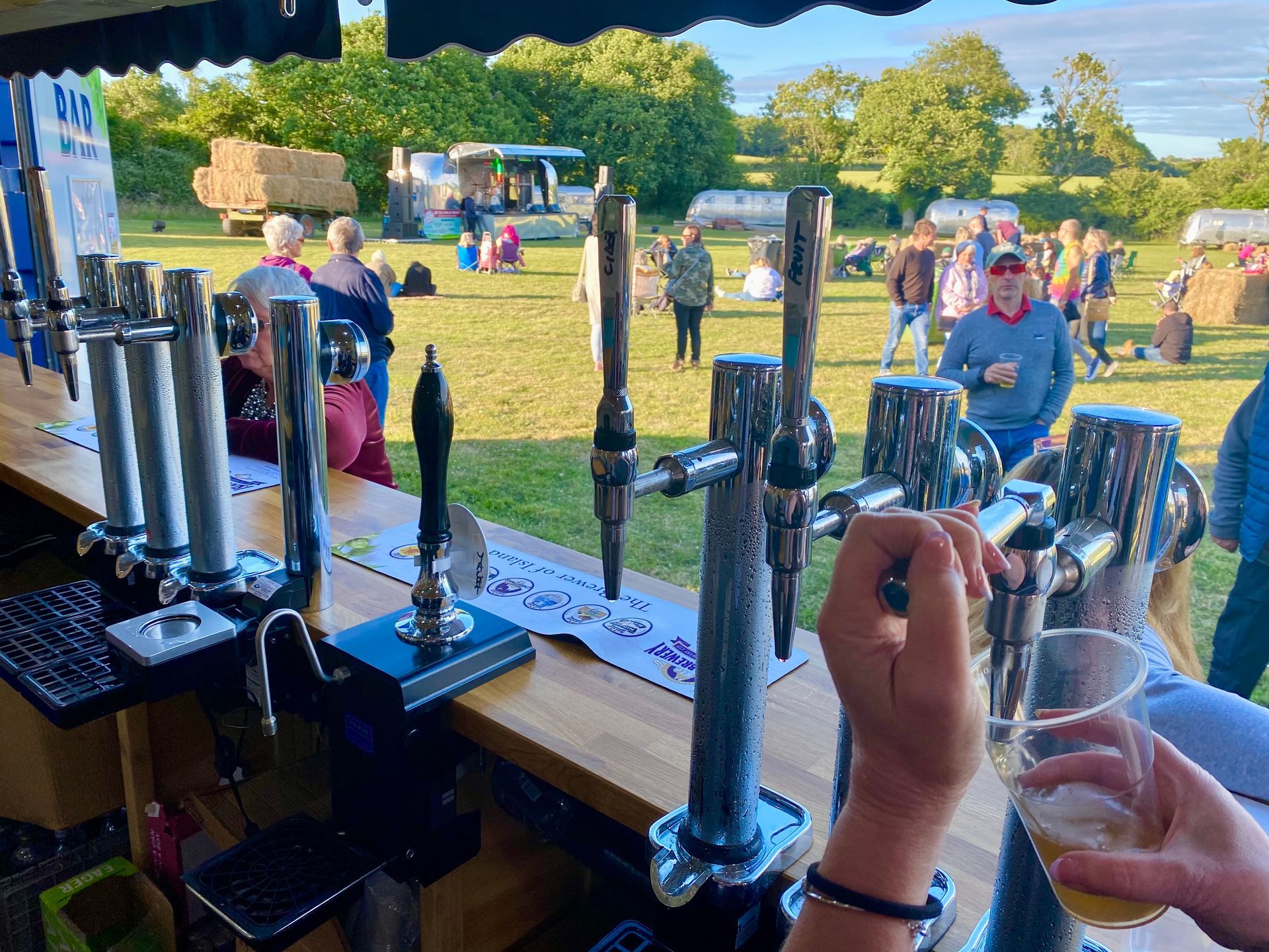 A person pours beer from a tap at an outdoor bar, with a crowd and field in the background.