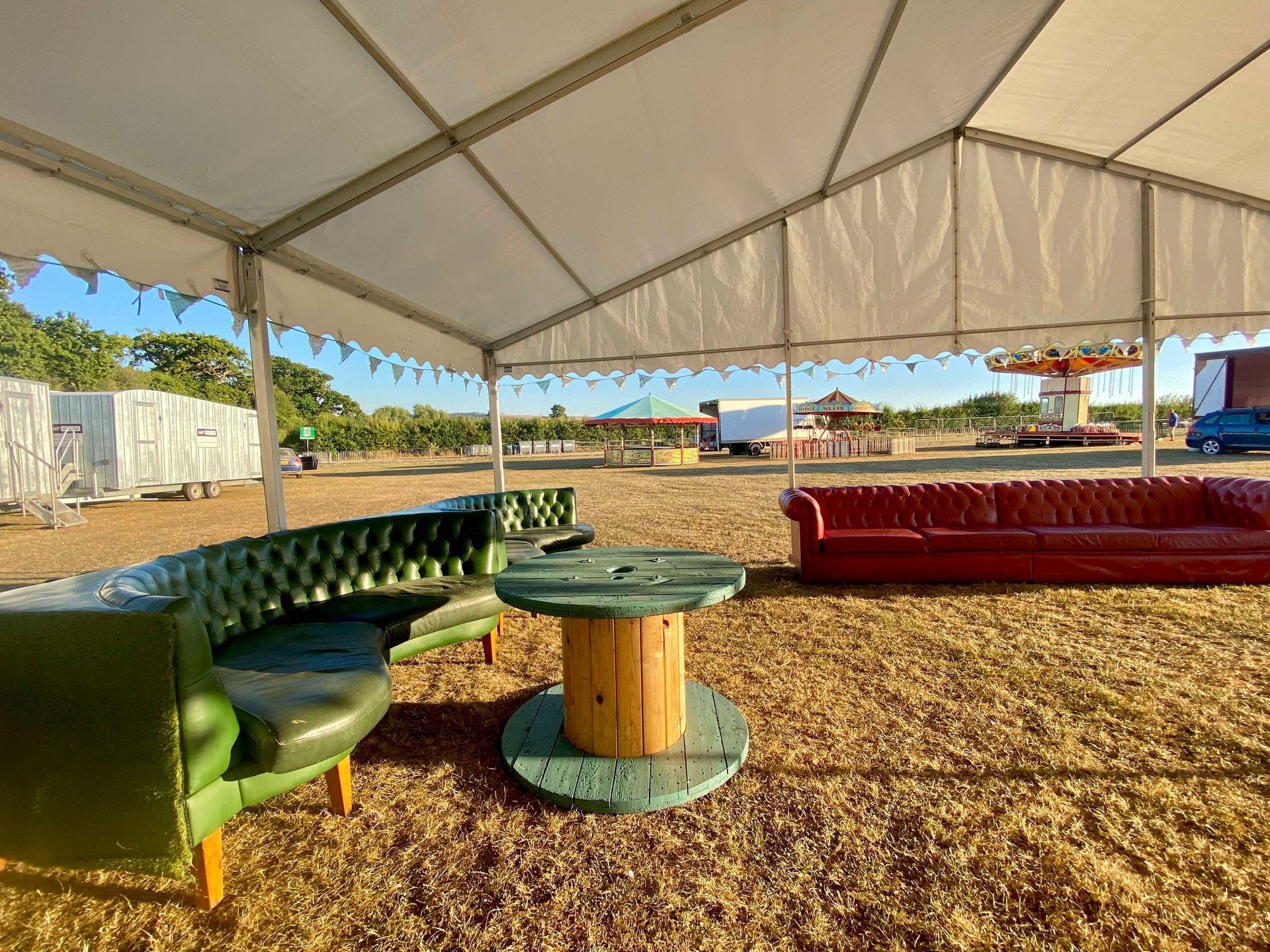 A tent with green and red Chesterfield sofas, a wooden spool table, and fairground in the background.