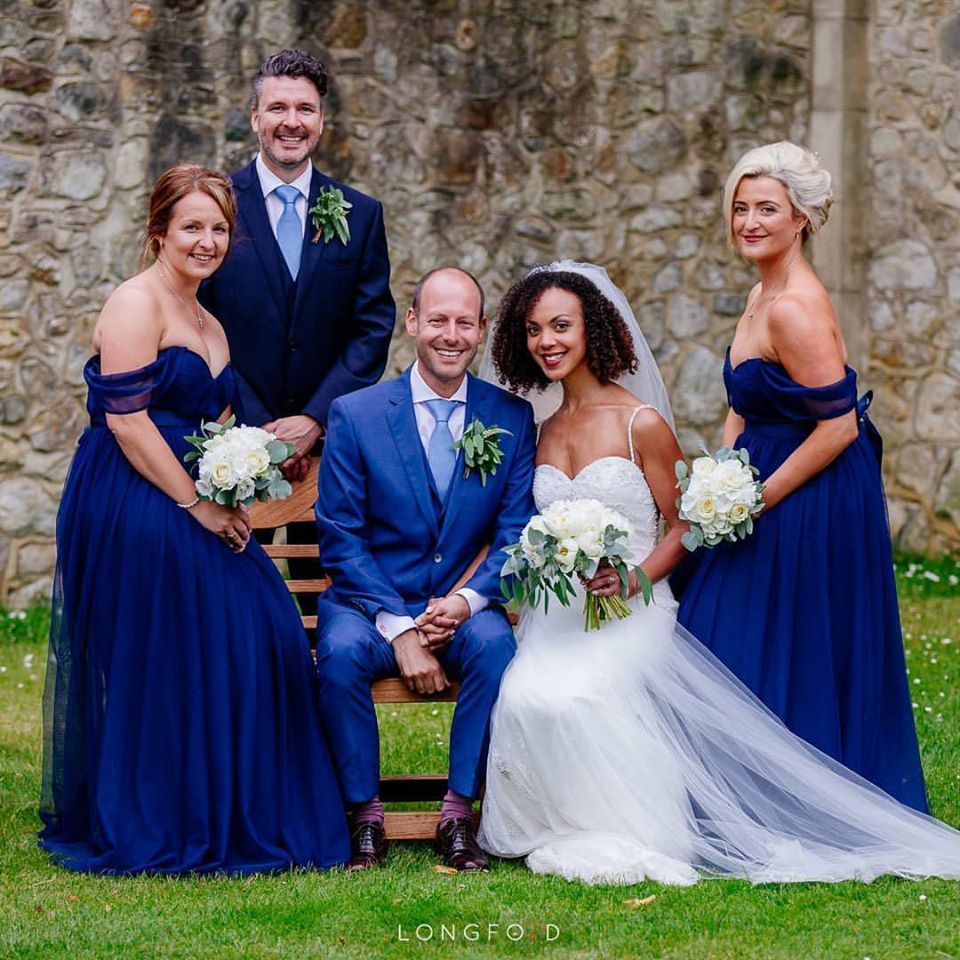 Wedding party posing in front of a stone wall. Bride and groom seated, flanked by bridesmaids and a groomsman. Navy blue dresses and suits.