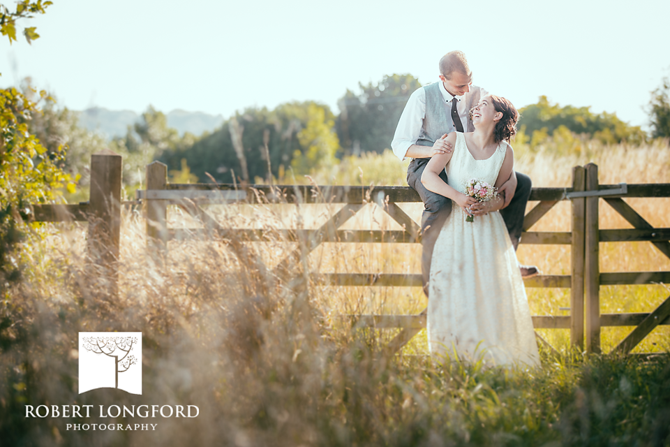 Newlyweds on a fence, golden-lit field. Groom sits; bride smiles, holding bouquet.