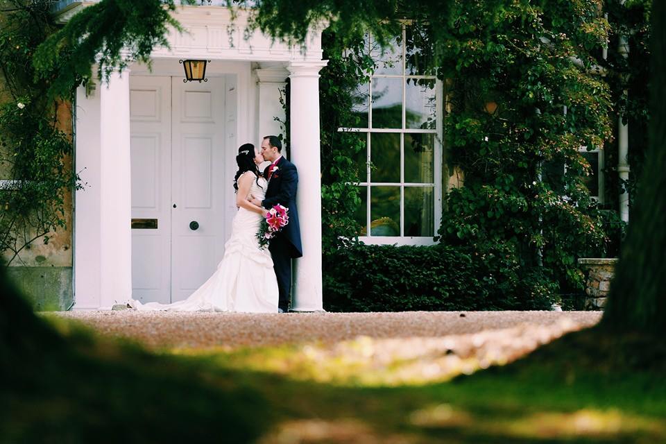 Bride and groom embracing in front of a white building with columns, greenery, and trees.