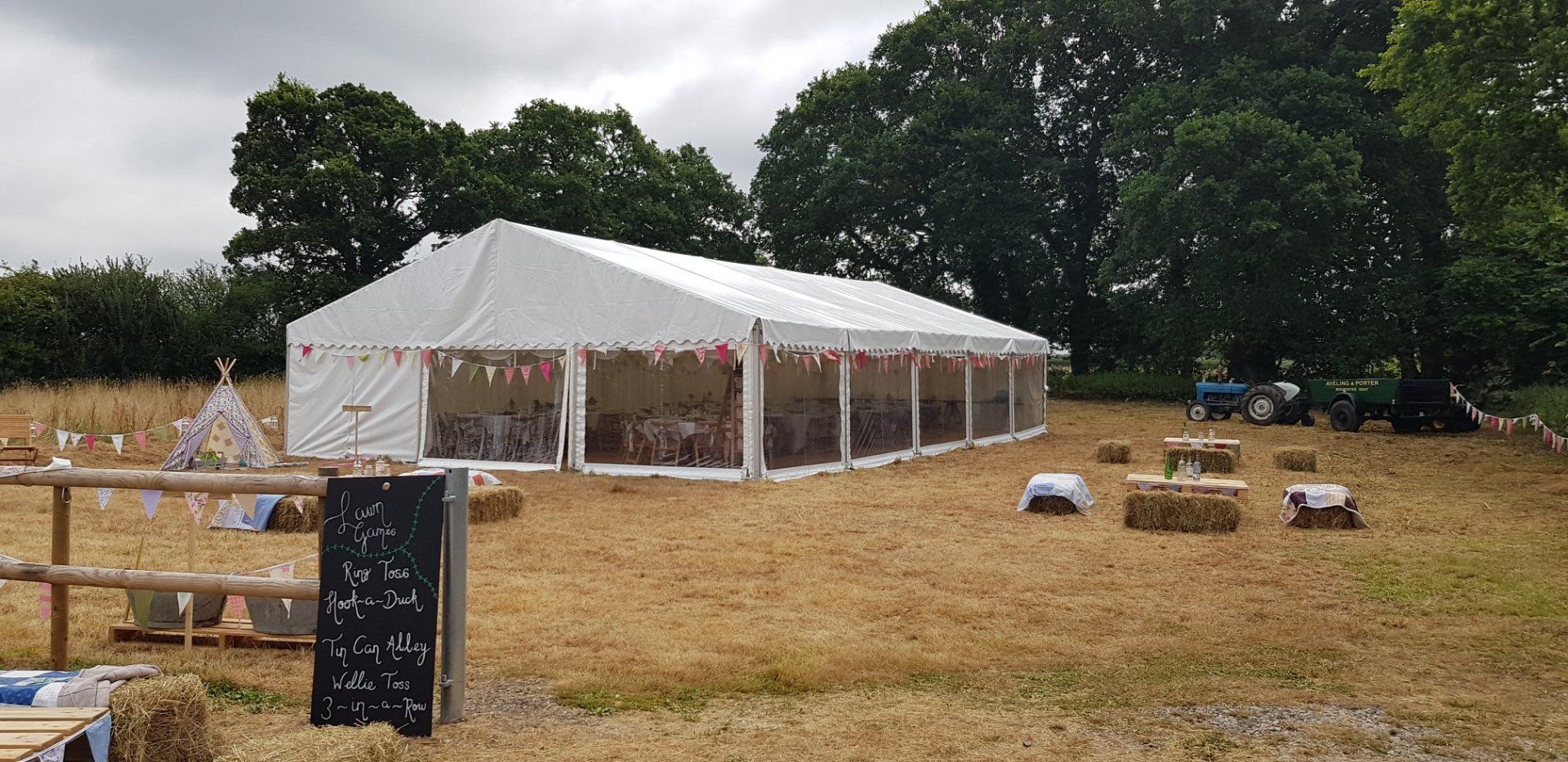 A white tent in a field with a wooden fence, hay bales, and a sign. Trees in the background.