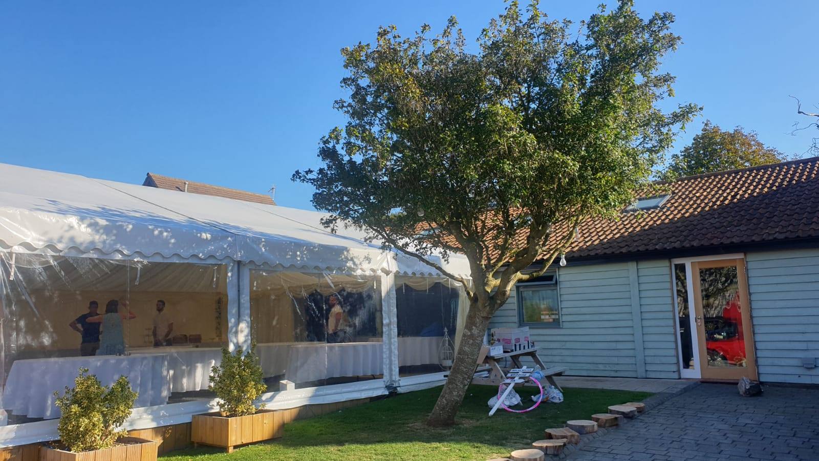 Outdoor event setup with a white tent, a building, and a tree on a sunny day.