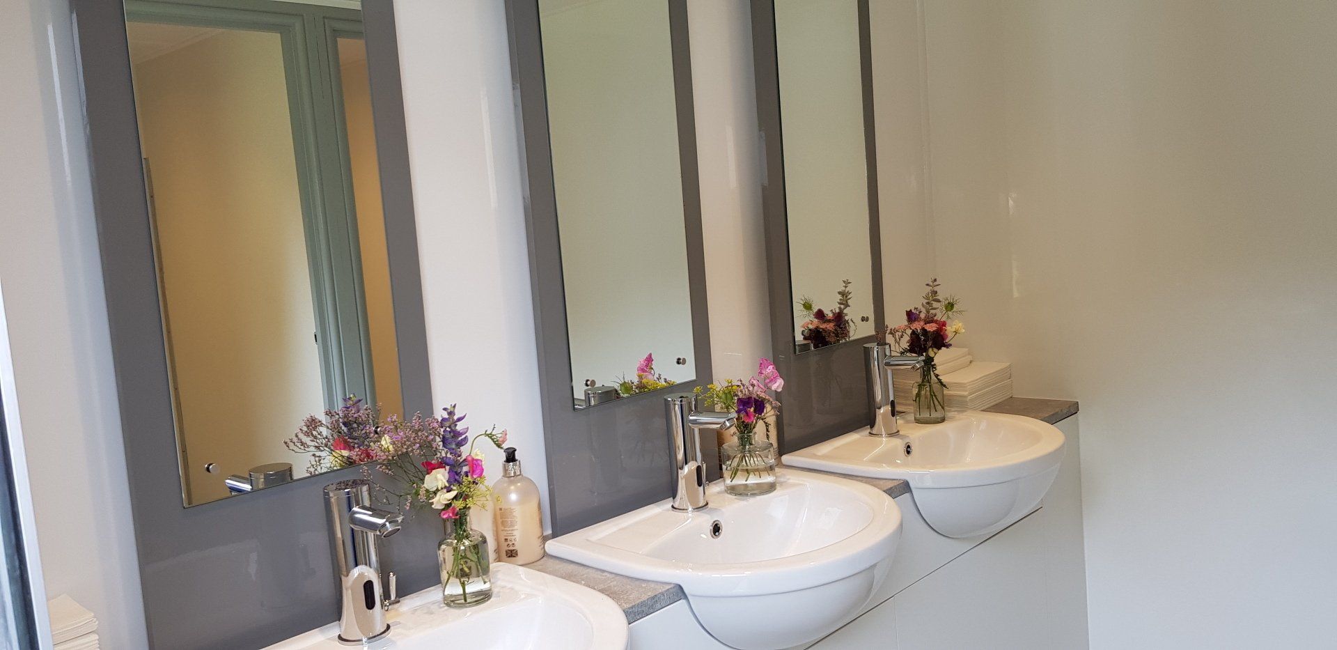 Three white sinks with mirrors, flowers, and soap bottles in a restroom.