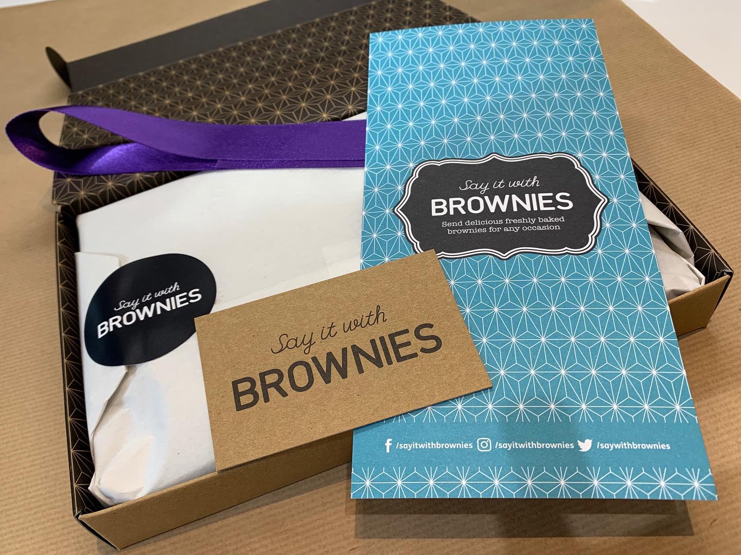 Open brownie box with purple ribbon, wrapped brownie package, brown card, and blue patterned sleeve.