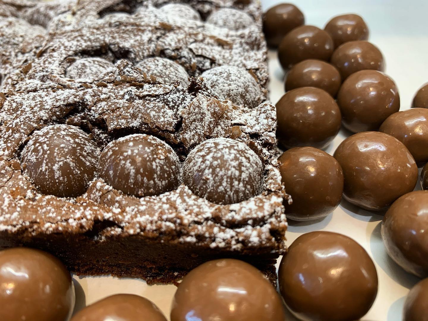 Brownie square topped with Maltesers and dusted with powdered sugar, surrounded by Maltesers.