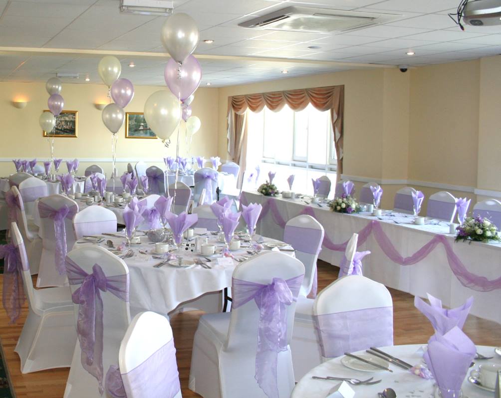 Wedding reception room set for a party, decorated in white and lavender, with balloons and table settings.