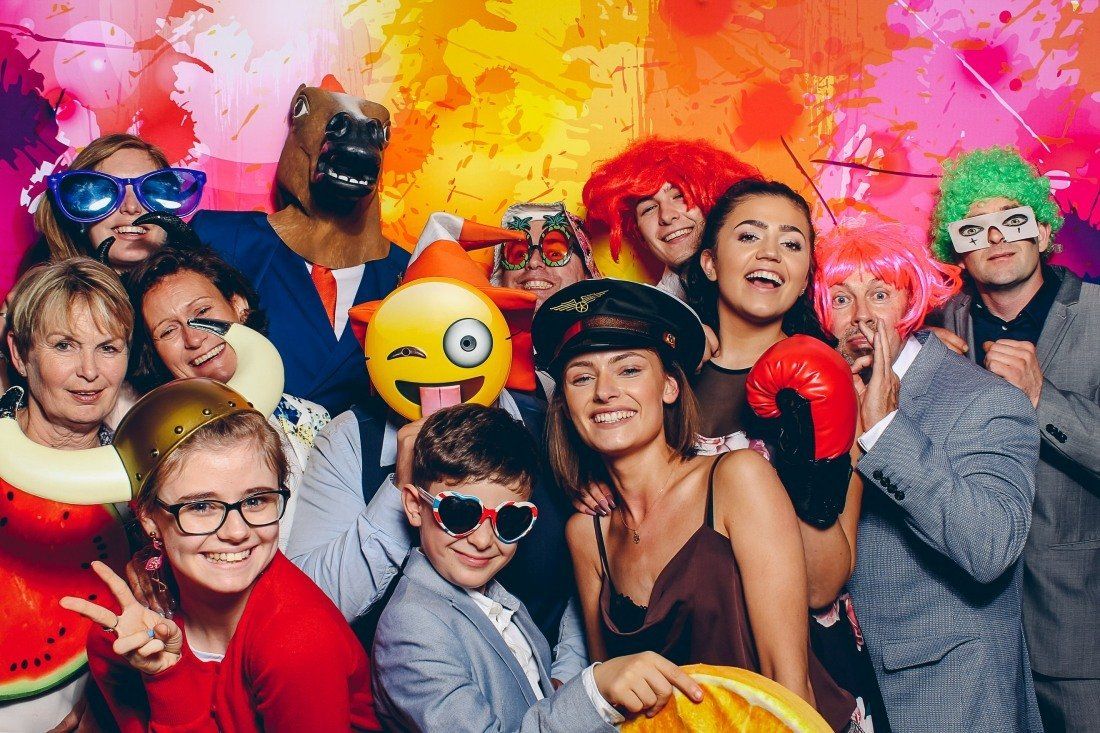 Group of people posing with props at a party, including wigs, masks, and sunglasses. Colorful backdrop.