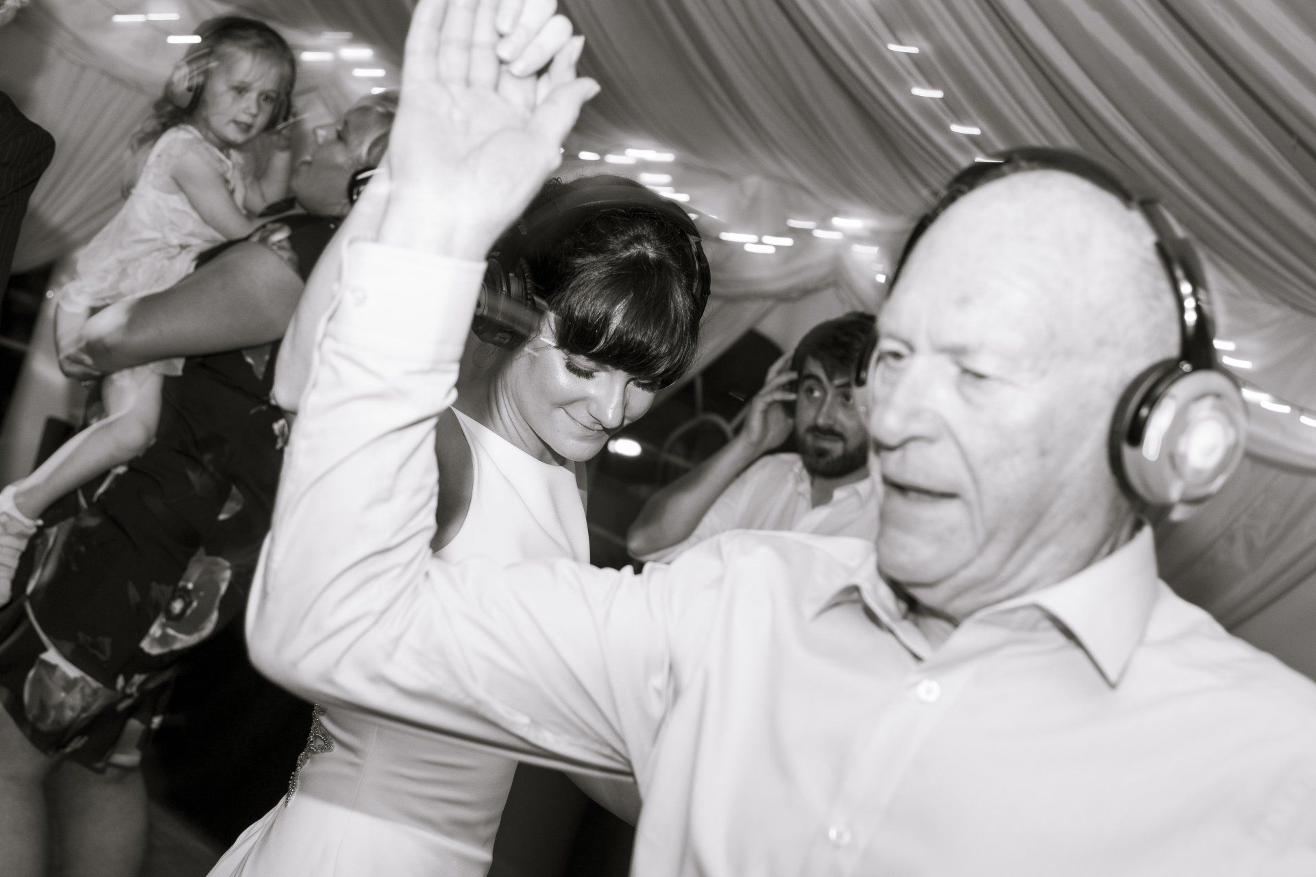 Man with headphones dances, arm raised. Woman dances beside him. Others in background.
