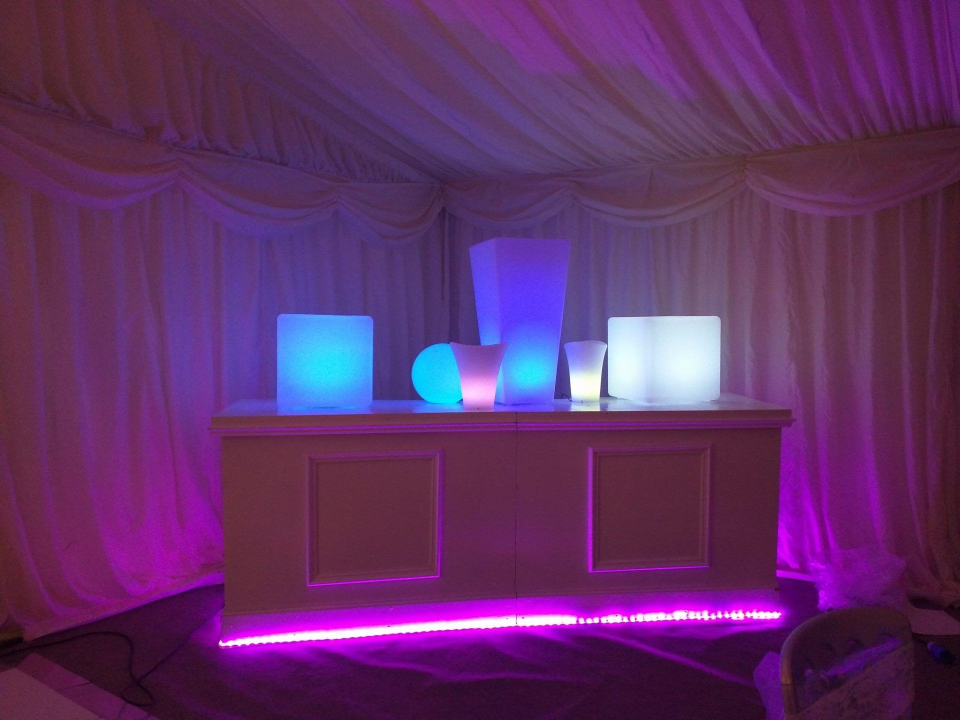 Bar with glowing white and blue illuminated decorations inside a white tent, lit by purple lights.