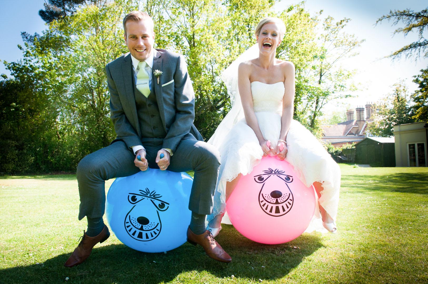Bride and groom on space hoppers, outside on lawn, both smiling. The groom is in gray suit, bride in white dress.