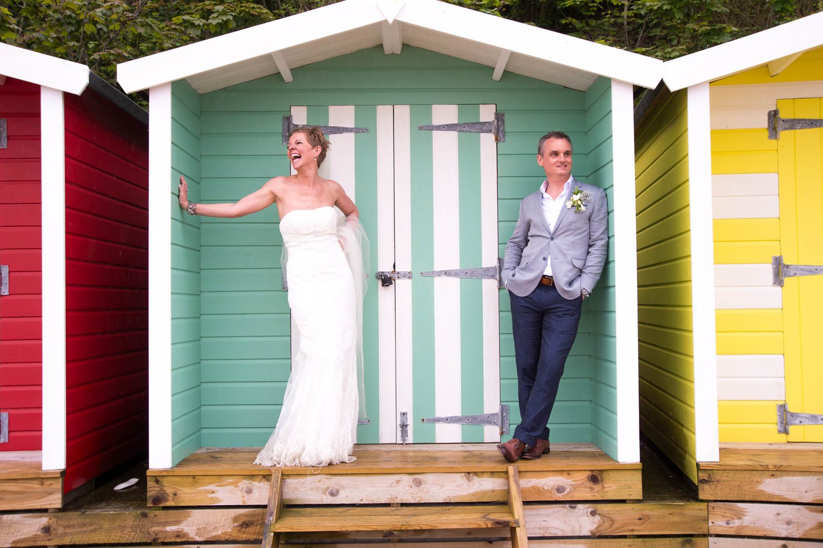 Bride and groom in wedding attire pose by a teal beach hut, other colorful huts alongside.