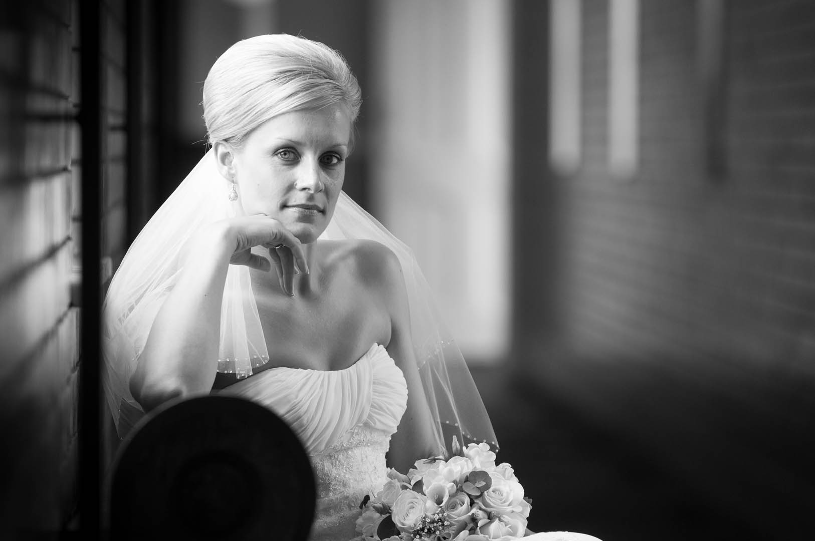 Bride in strapless gown rests hand on chin, looking at camera. Holds bouquet, seated by a brick wall and window.