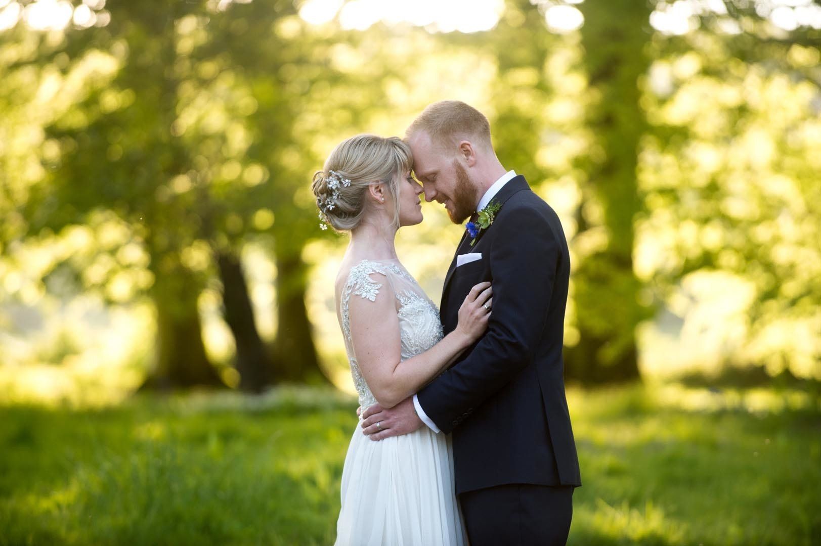 Bride and groom embrace outdoors in front of trees. Soft lighting, grassy area, smiling, close together.