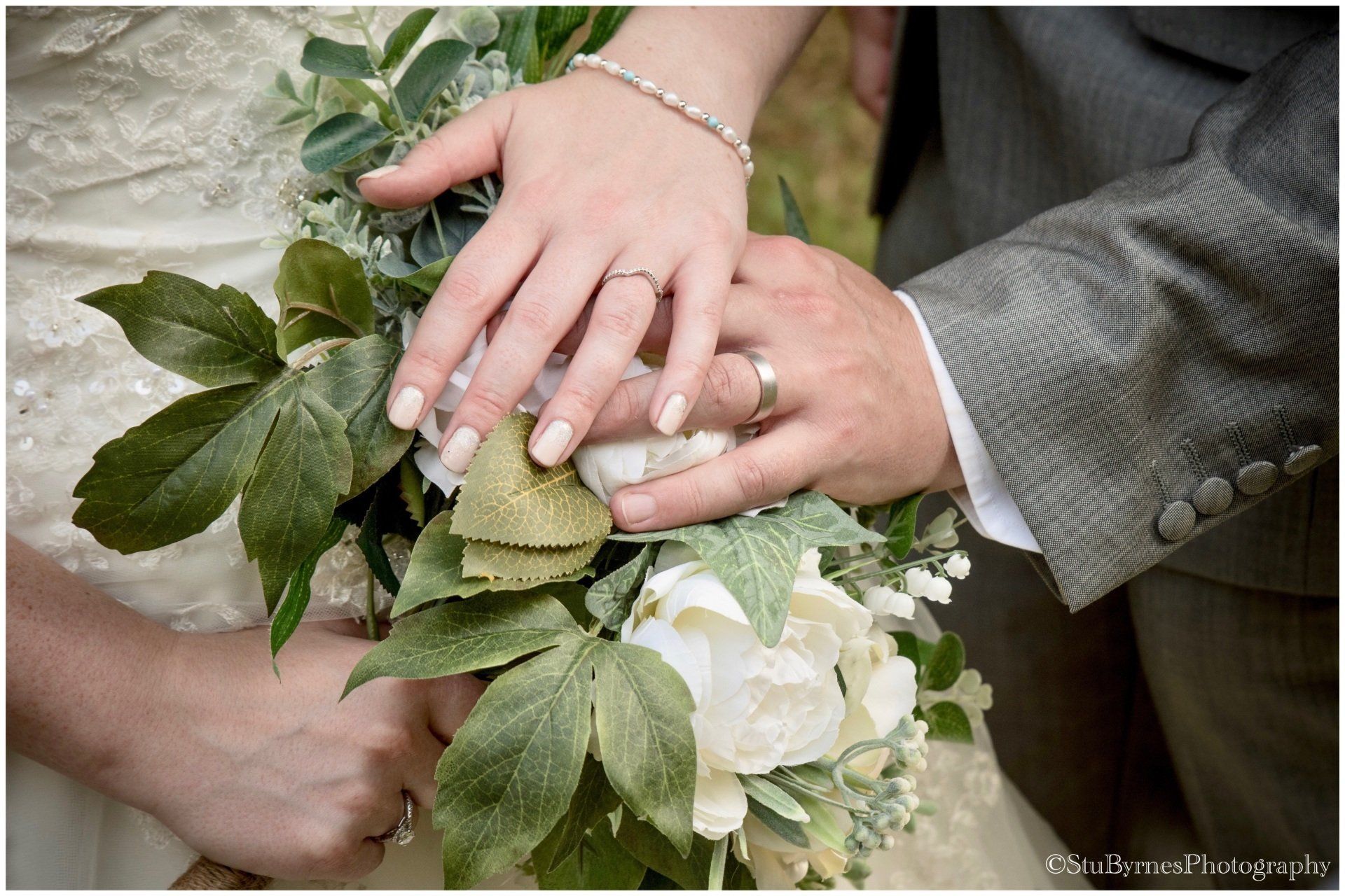Bride and groom's hands with wedding rings on a bouquet of white flowers and green leaves.