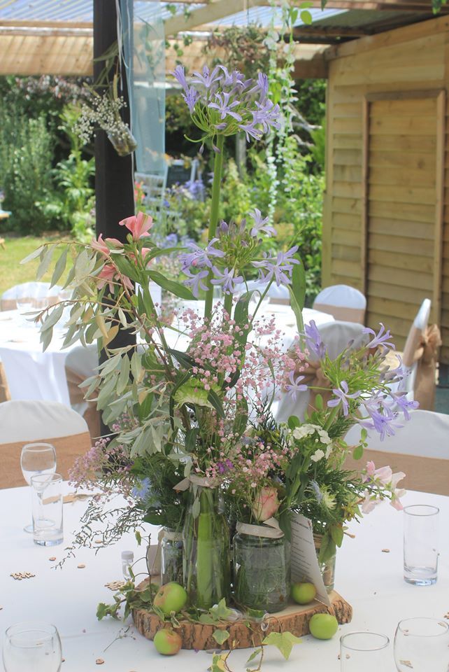 Floral centerpiece on table with clear glasses; green, pink, and purple flowers.
