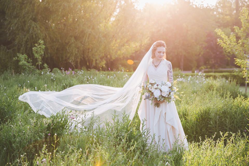 Bride in flowing gown with long veil, holding bouquet, standing in a field with sunlight.