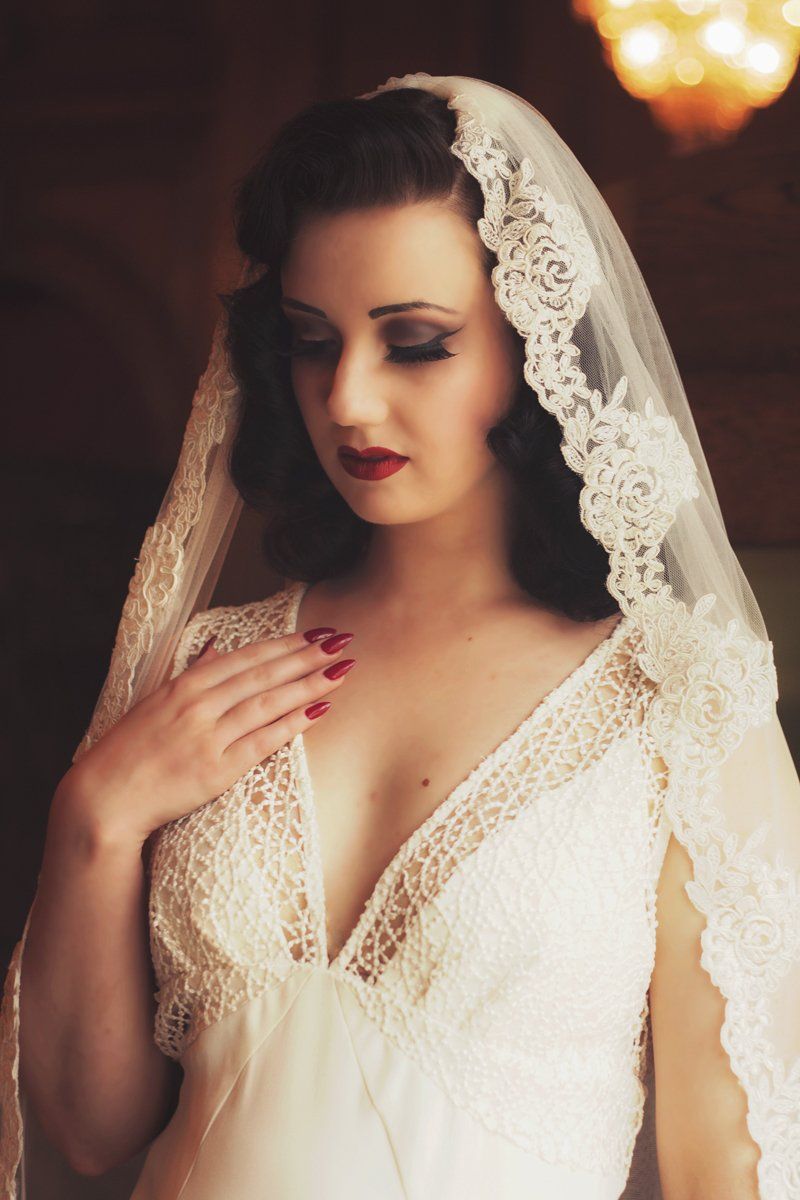 Woman in wedding dress with veil, hand on chest, dark hair, red lips, vintage setting.
