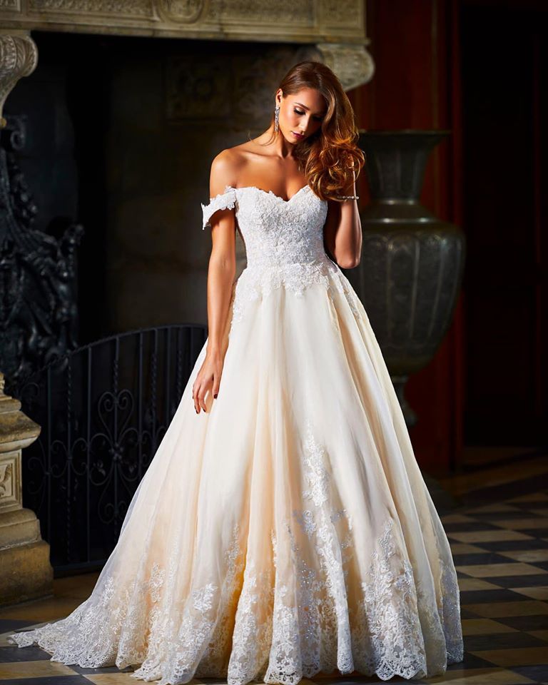 Woman in off-the-shoulder wedding gown, standing indoors. The dress is ivory with lace detailing, and she is gazing down.