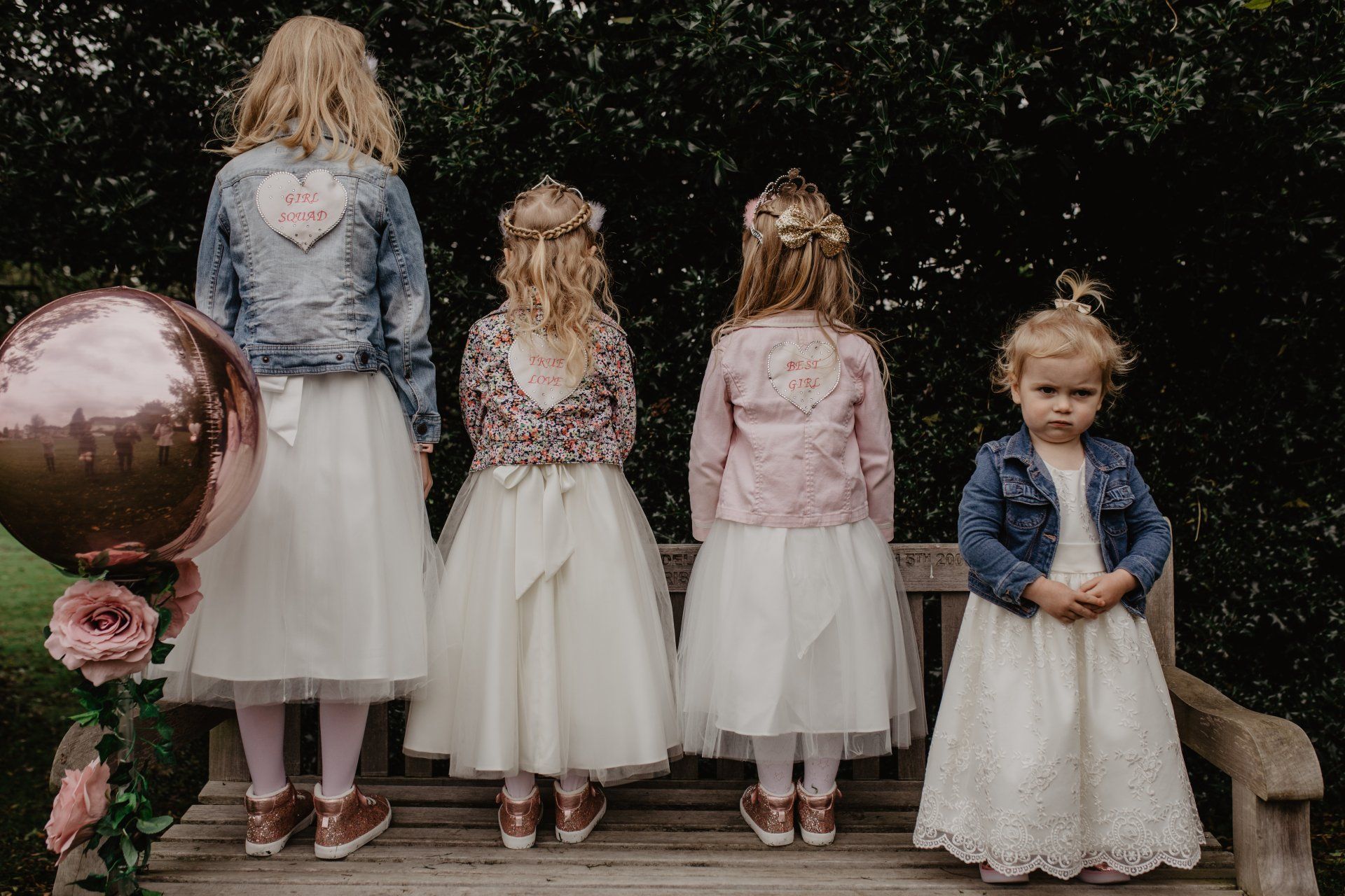 Four young girls, backs to the camera, in dresses and jackets, stand on a bench, one with an unhappy expression.