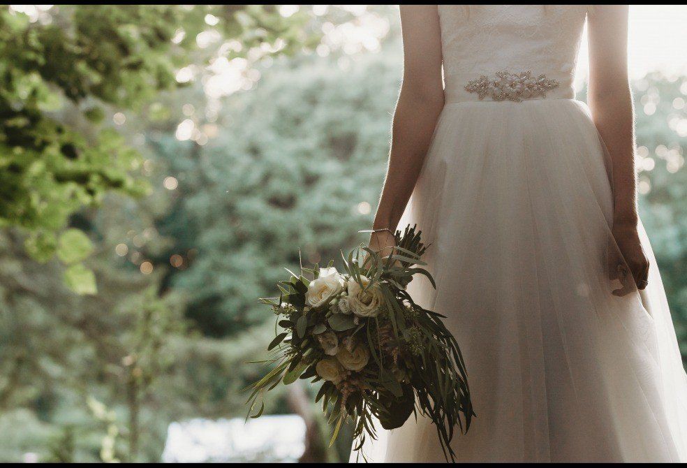 Bride in white dress holding bouquet, backlit by sunlight in a garden.