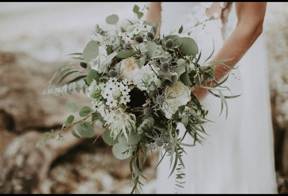 Bride holding a lush, natural bouquet with white and green flowers and eucalyptus; outdoor setting.