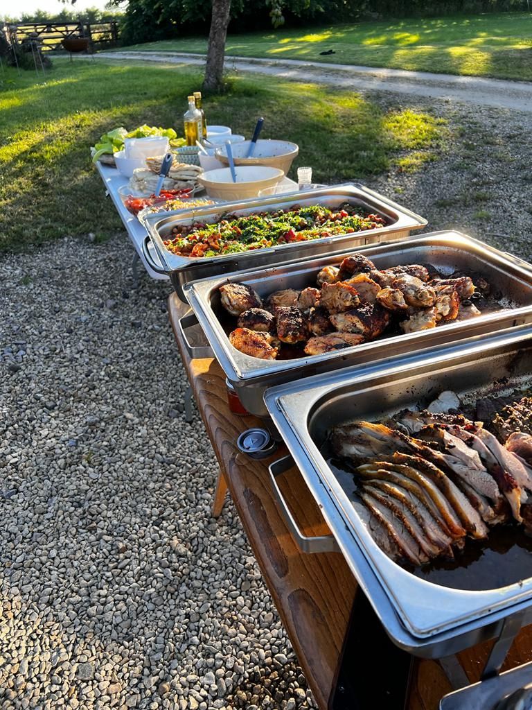 Outdoor buffet with various food trays on a table in a gravel yard, sunny.