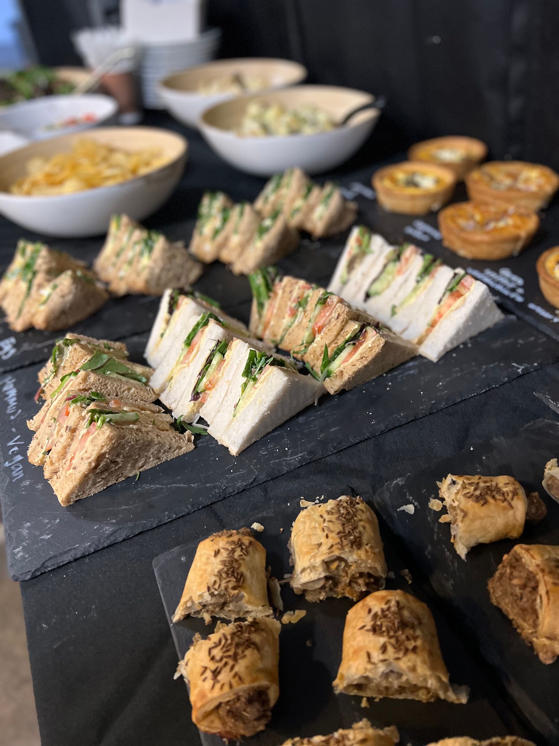 Assortment of sandwiches, quiches, and sausage rolls on black slate platters; bowls of salad and chips in background.