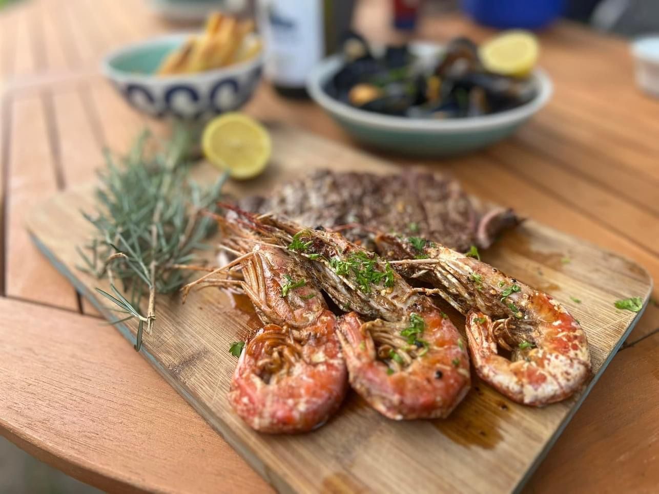 Grilled shrimp, steak, and mussels on a wooden board with rosemary and lemon, on a table.