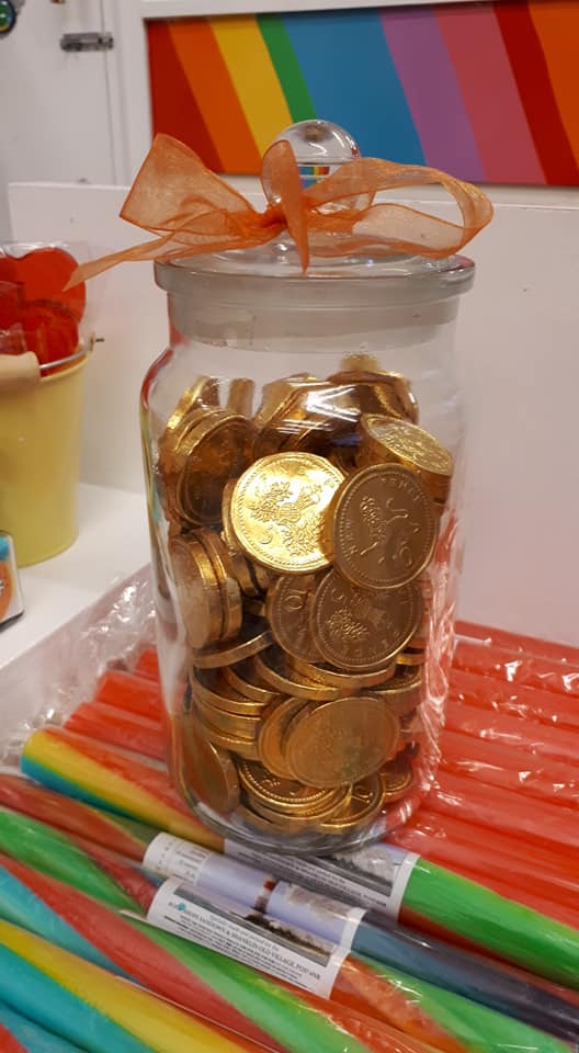 Glass jar filled with gold chocolate coins, tied with orange ribbon. Rainbow candy sticks below.
