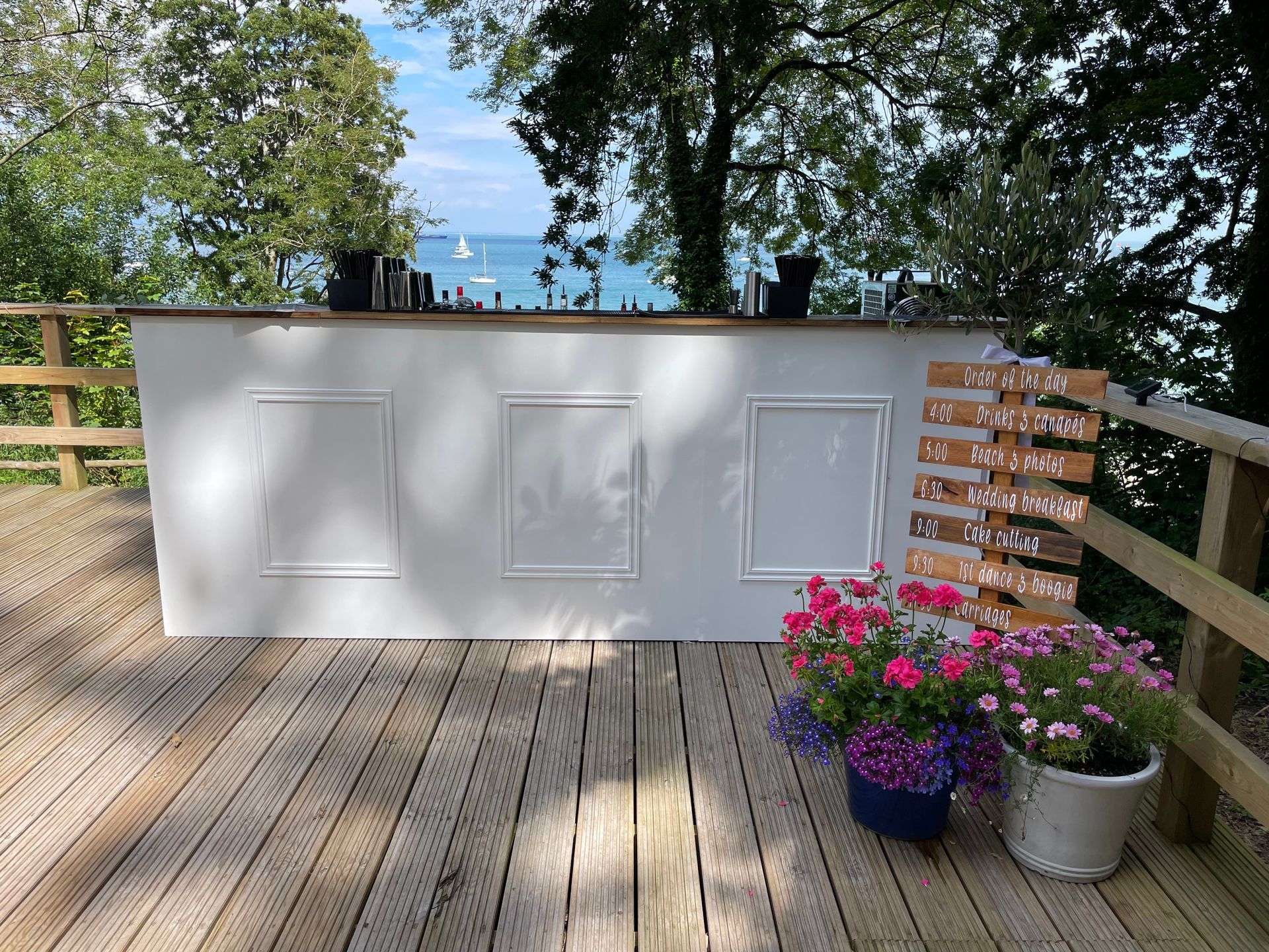 White bar on a wooden deck with framed panels and floral arrangements, overlooking a seascape.