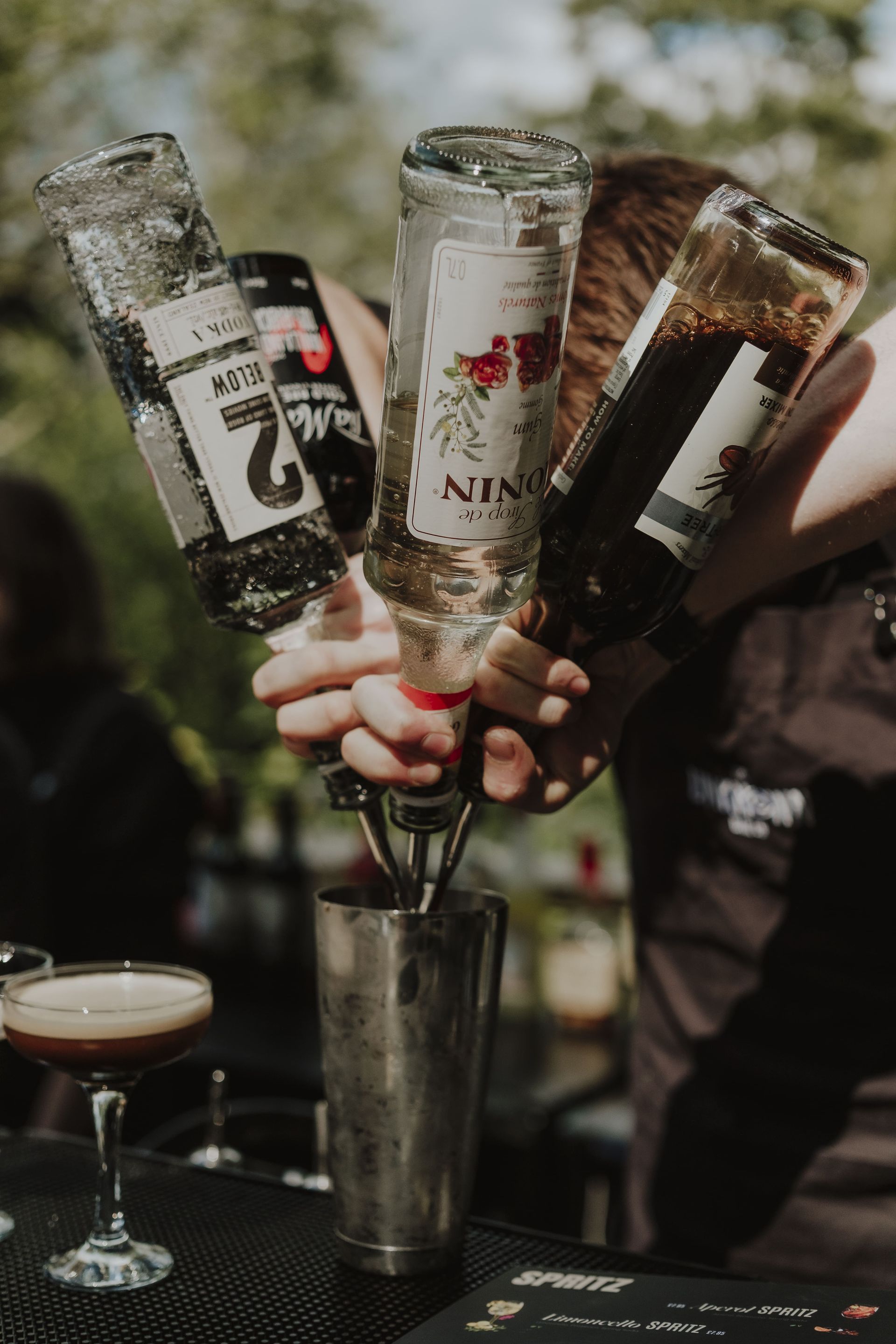 Bartender pouring spirits from three bottles into a shaker, cocktail on bar.