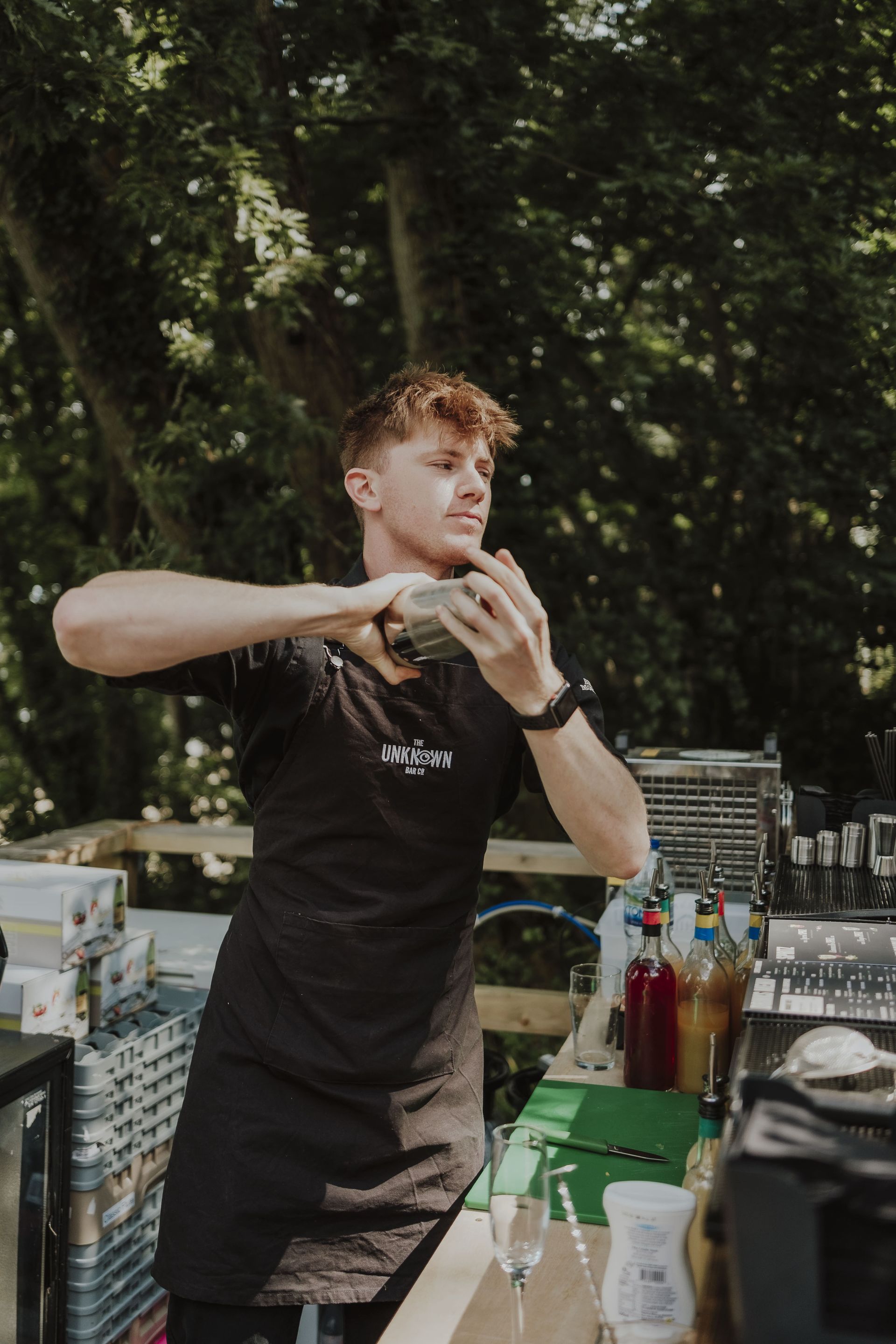 Bartender shaking a cocktail shaker outdoors; green bar with bottles and a tall tree background.