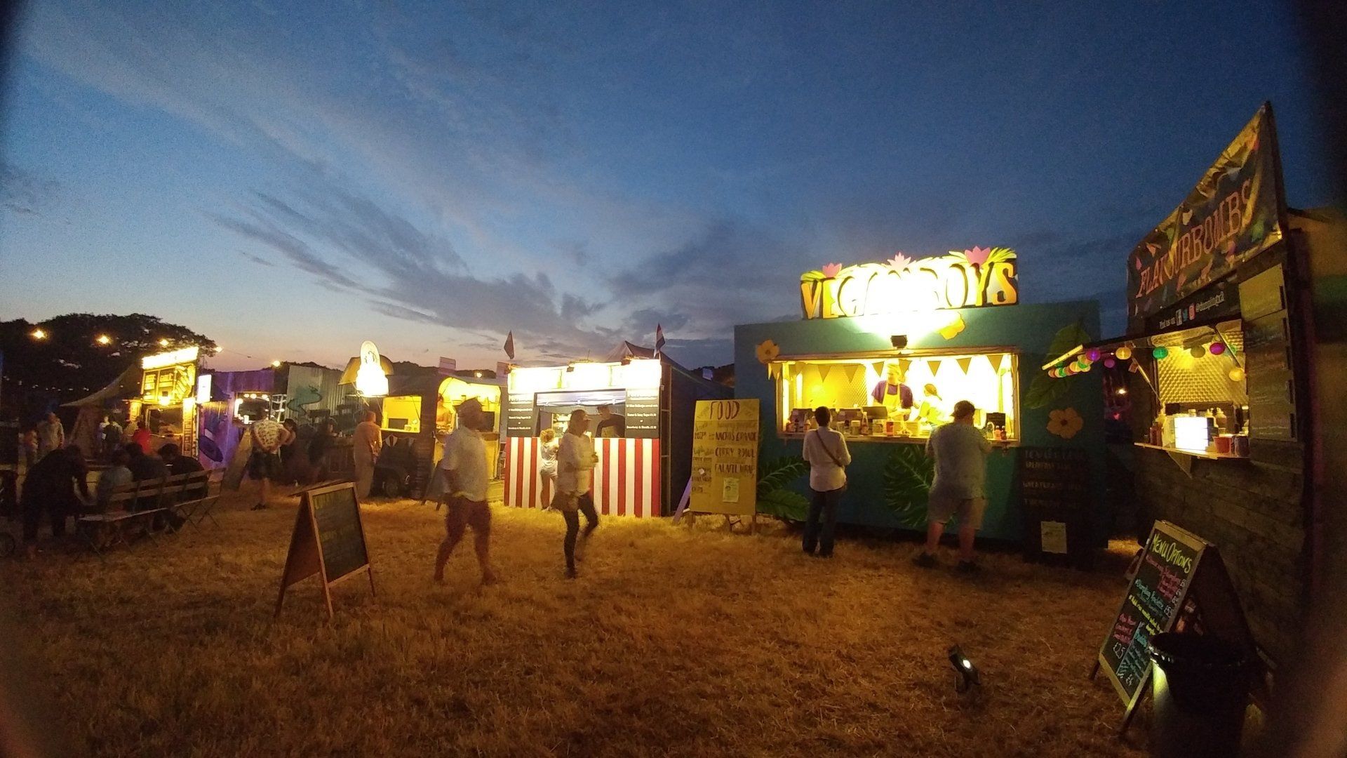 Food stalls at dusk, with people milling about. Illuminated signs, colorful food trucks, grassy field, and a dark blue sky.