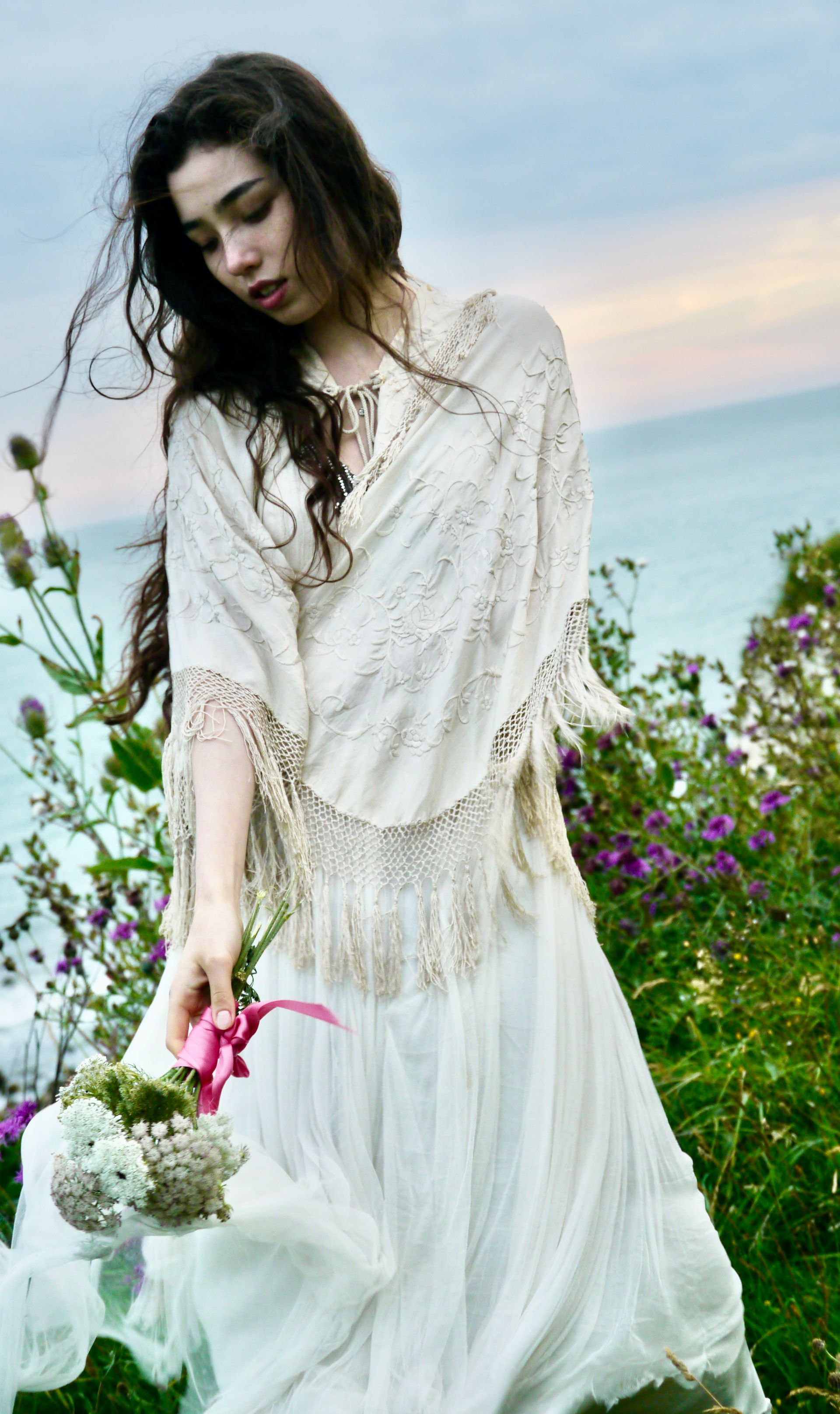 Woman in white dress by the ocean, holding flowers, looking down.
