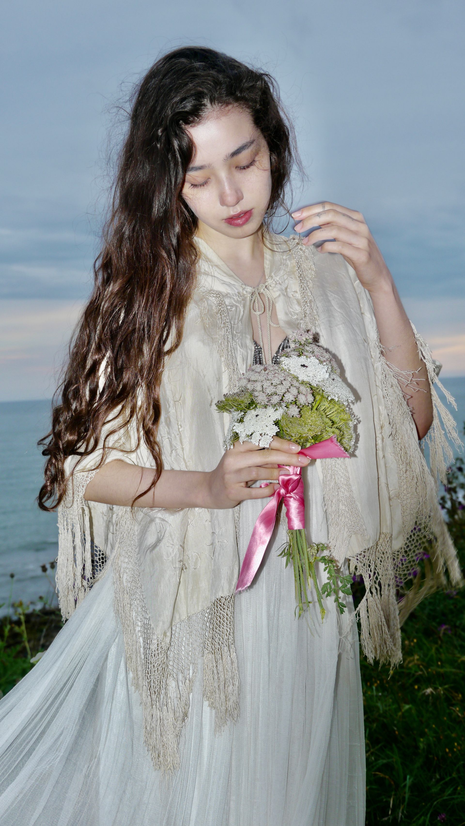 Woman holding flowers by the ocean, wearing a white dress and lace shawl.