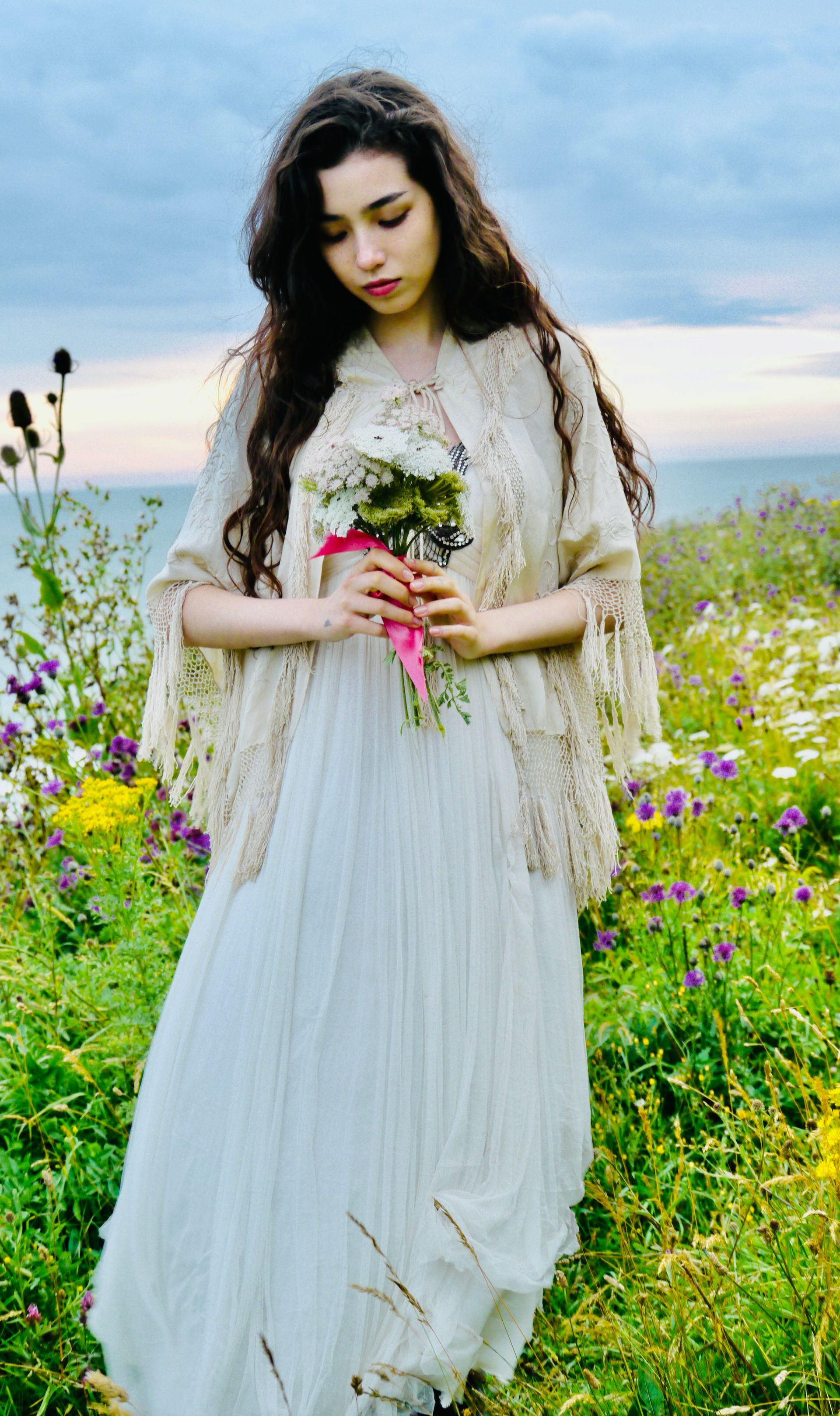 Woman in white dress and shawl holding flowers in a field of wildflowers.