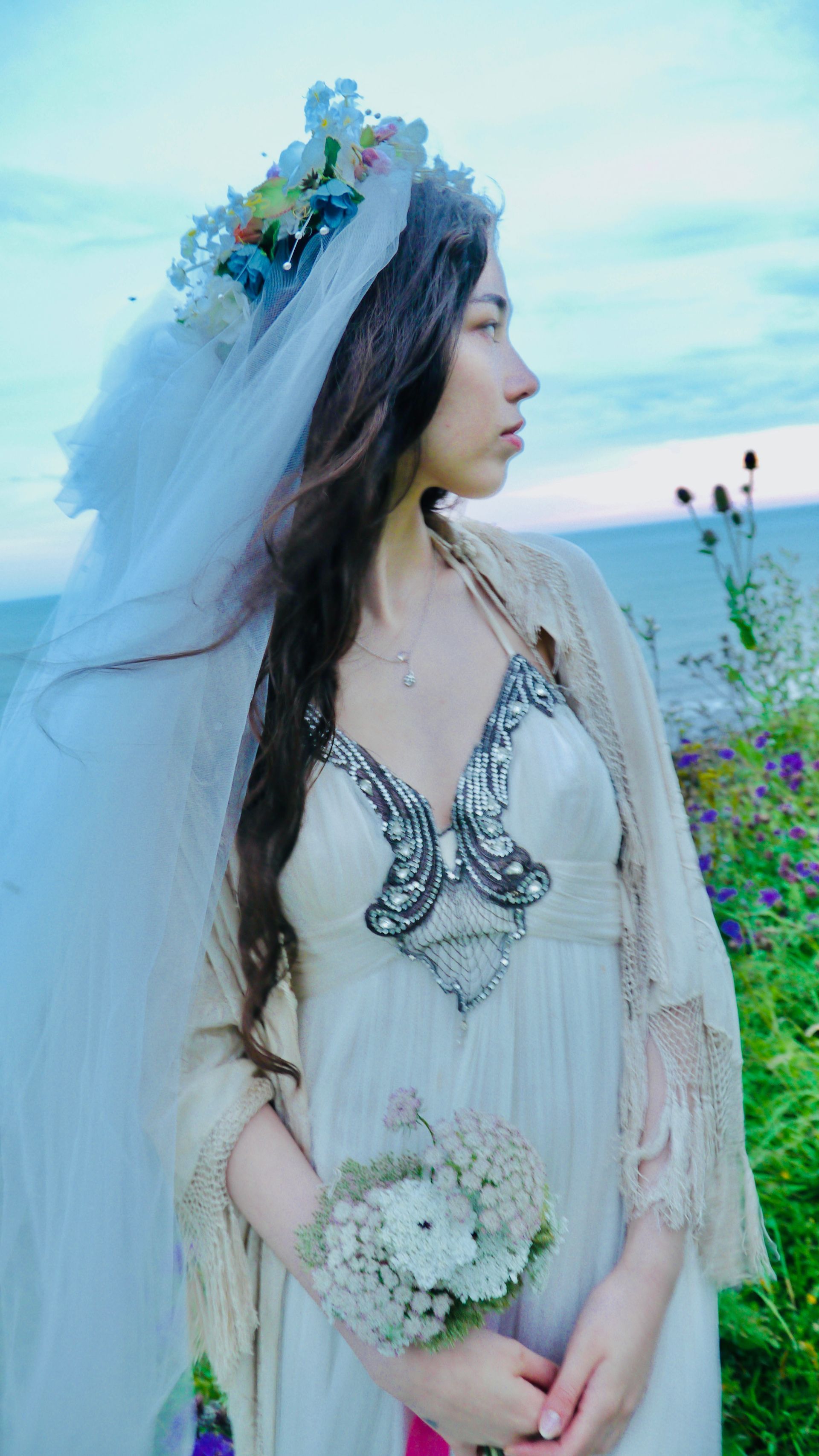 Woman in white dress and floral headpiece, holding bouquet, looking toward ocean.