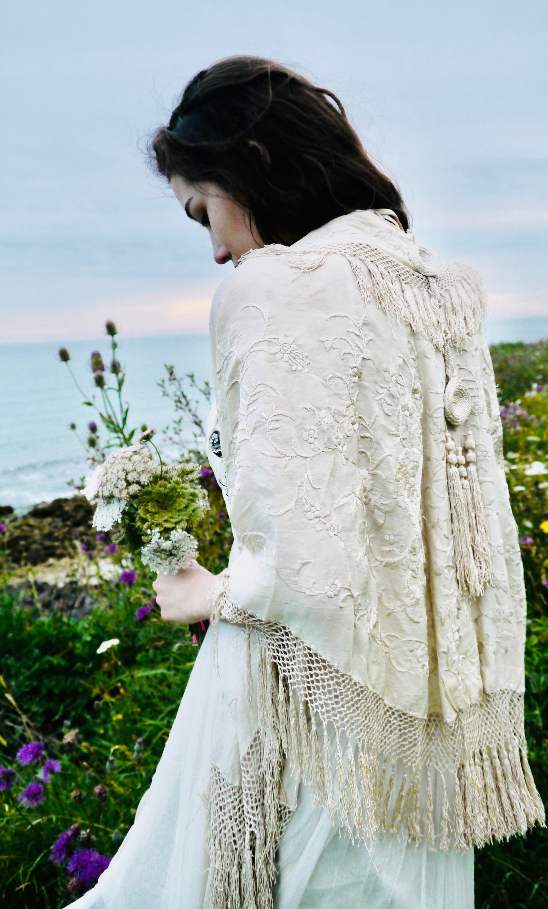 Woman in a lace shawl holding flowers, walking near the ocean and wildflowers.