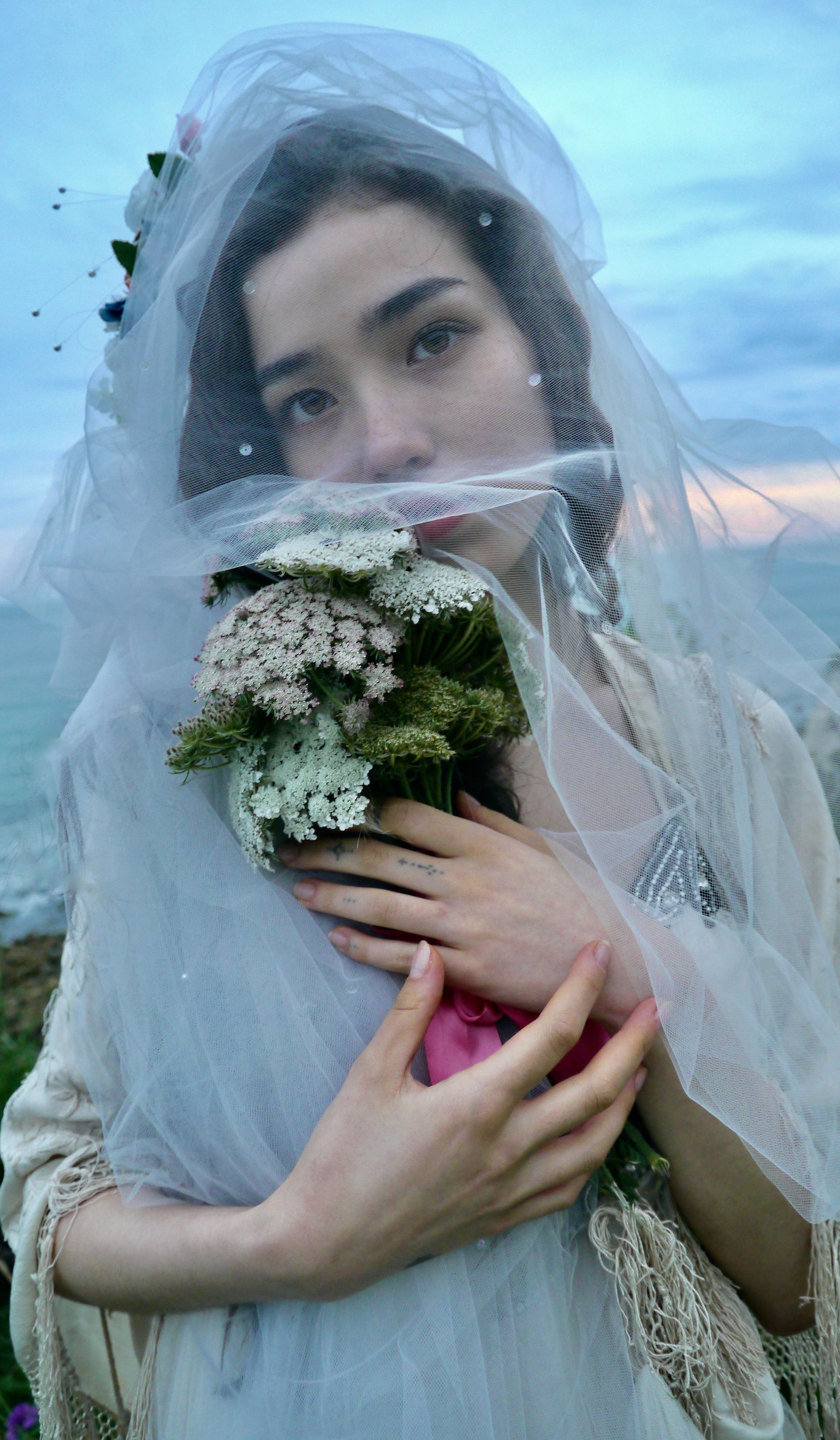 Woman holding bouquet, partially veiled, by the ocean.