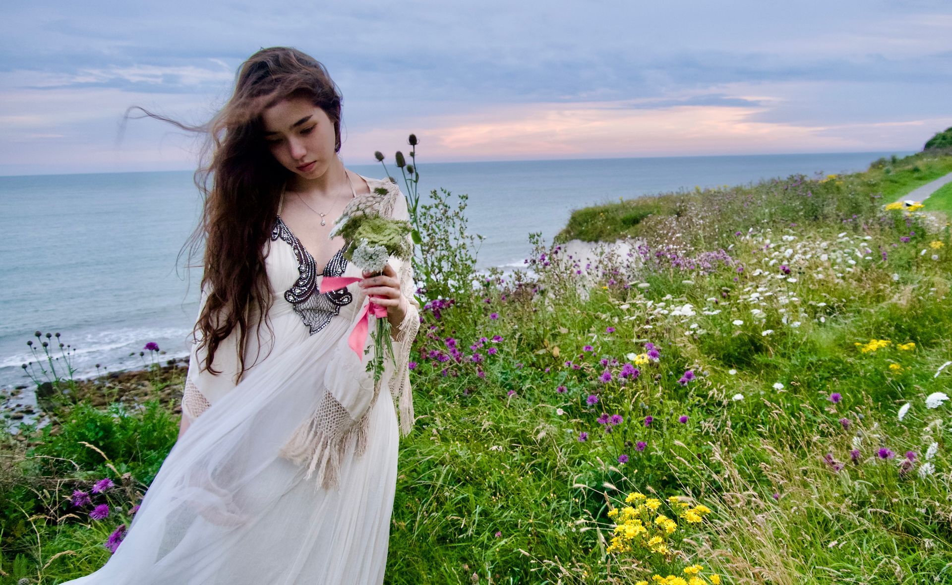Woman in white dress holding wildflowers by the ocean.
