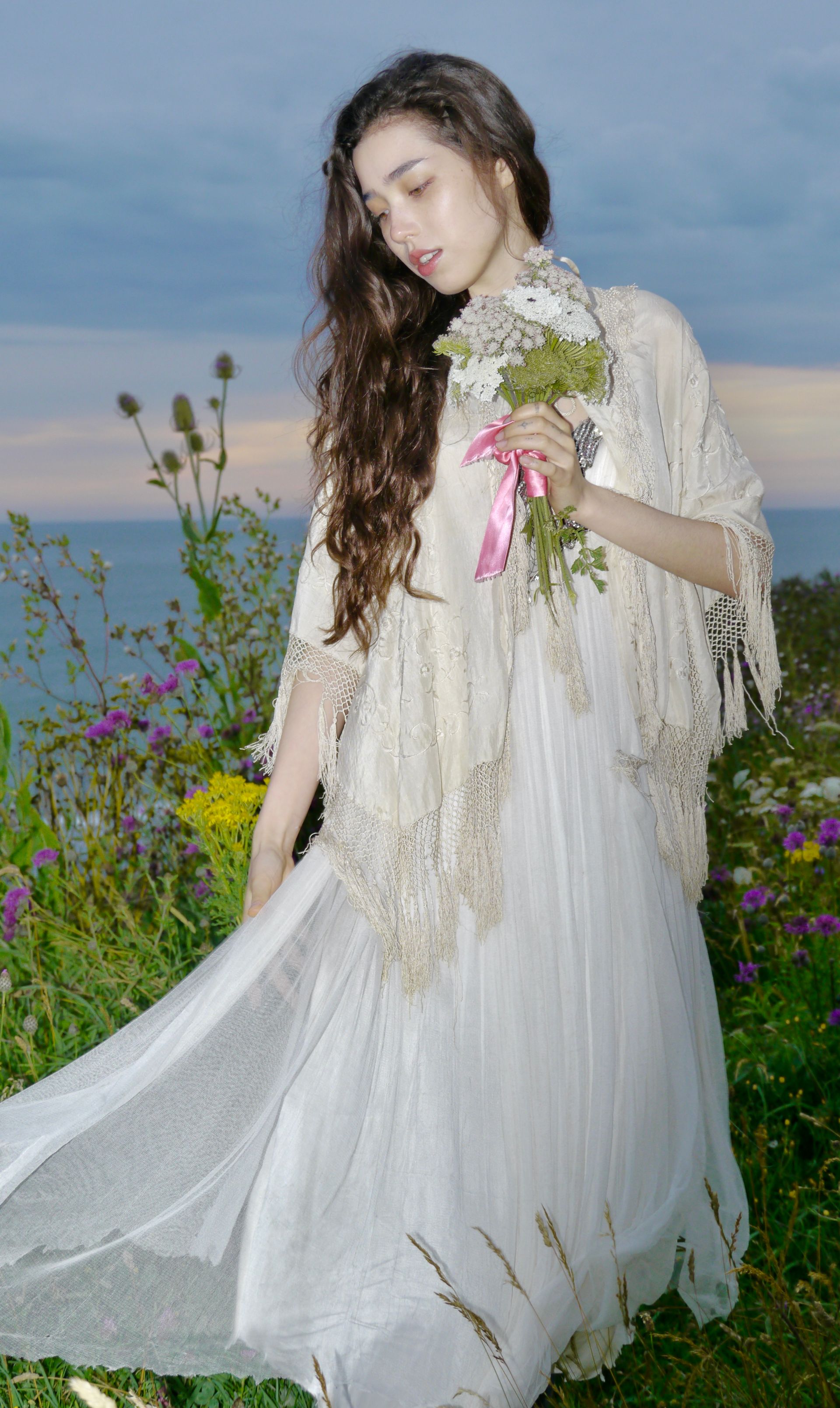 Woman in white dress and shawl holding flowers outdoors near water.