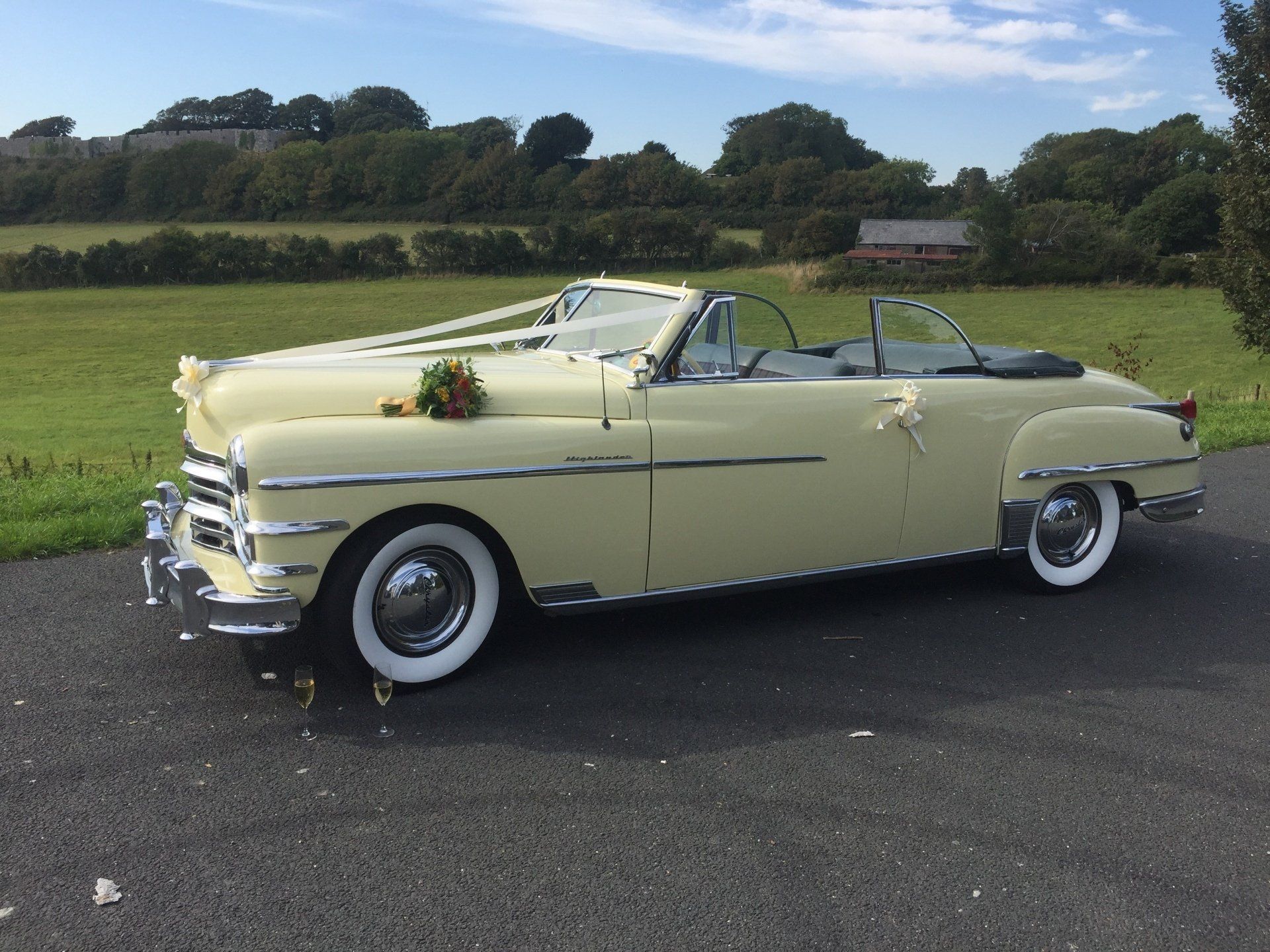 Classic yellow convertible decorated for a wedding, parked on a paved road, greenery in the background.