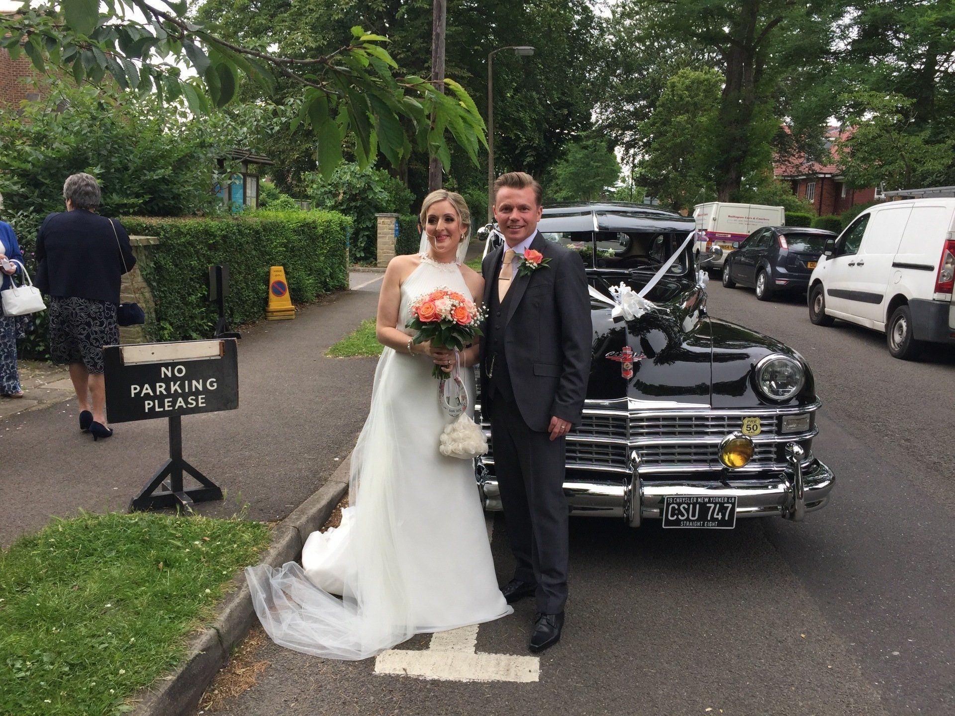 Bride and groom pose by a black vintage car. The bride wears a white gown and holds flowers. The groom wears a gray suit.
