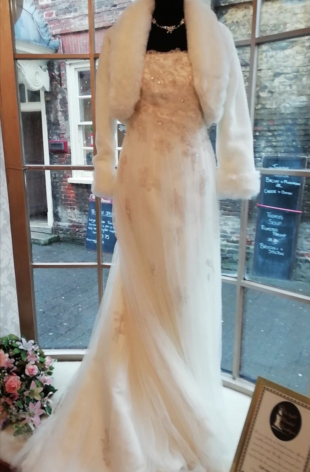 Wedding dress on a mannequin in a window, with a faux fur shrug, cream-colored with subtle golden detailing.