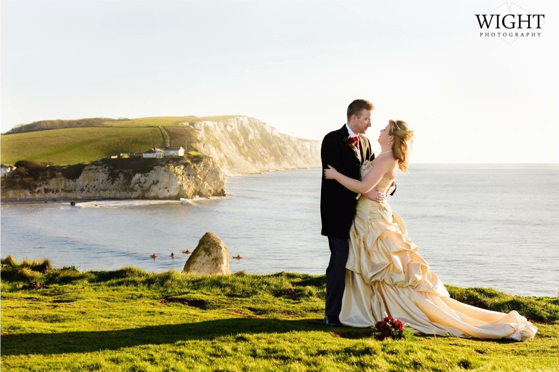 Couple embracing on a grassy cliff overlooking ocean and coastline. Bride in golden gown, groom in suit.