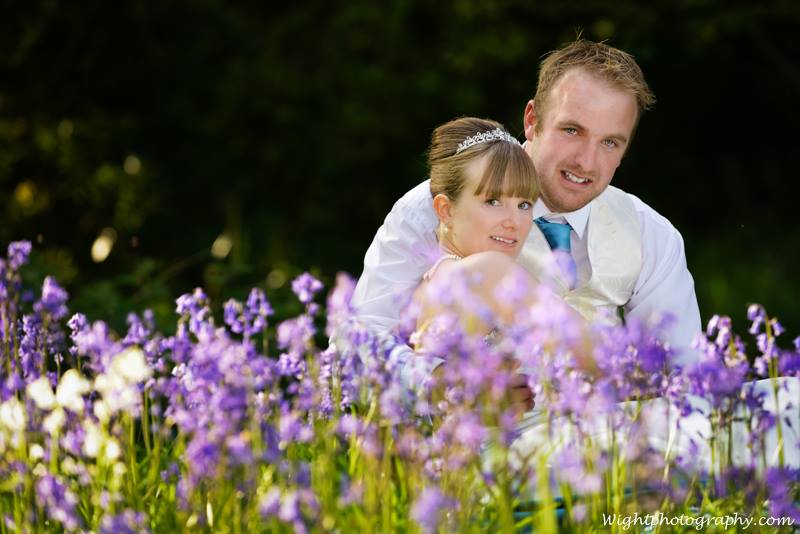 Couple embraces in field of purple flowers; man smiles, woman looks over shoulder.