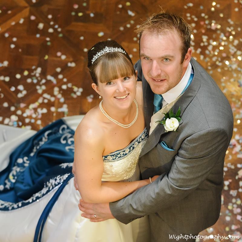 Bride and groom embrace on wooden floor, confetti scattered. Bride wears a white strapless gown with blue detailing; groom, a gray suit.