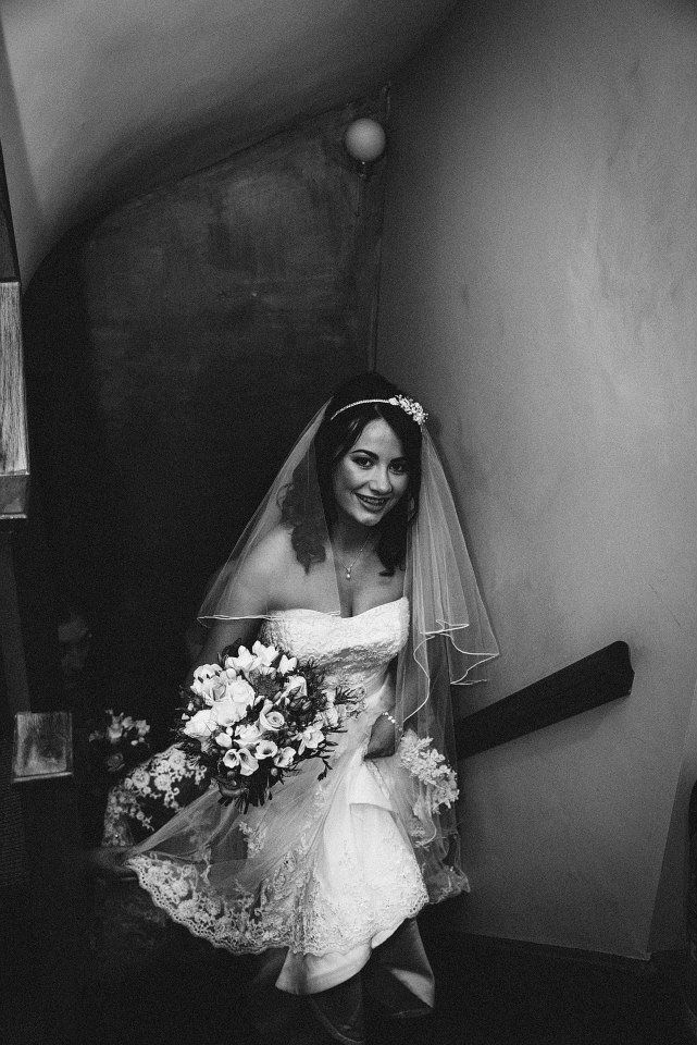 Bride in a strapless gown and veil, smiling as she descends a staircase, carrying flowers. Black and white.